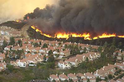 A cloud of black smoke and a line of fire loom over a town of homes with tan roofs. 